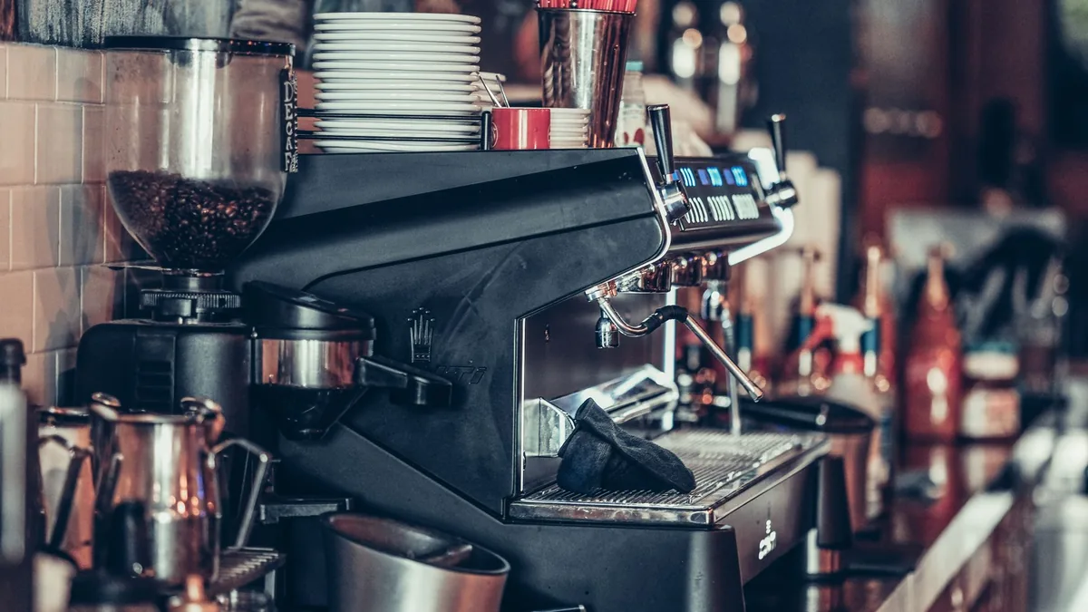 Máquina de espresso profesional en la barra de una cafetería, con la cesta del portafiltros a la vista, jarras de leche y un molinillo con granos al fondo.