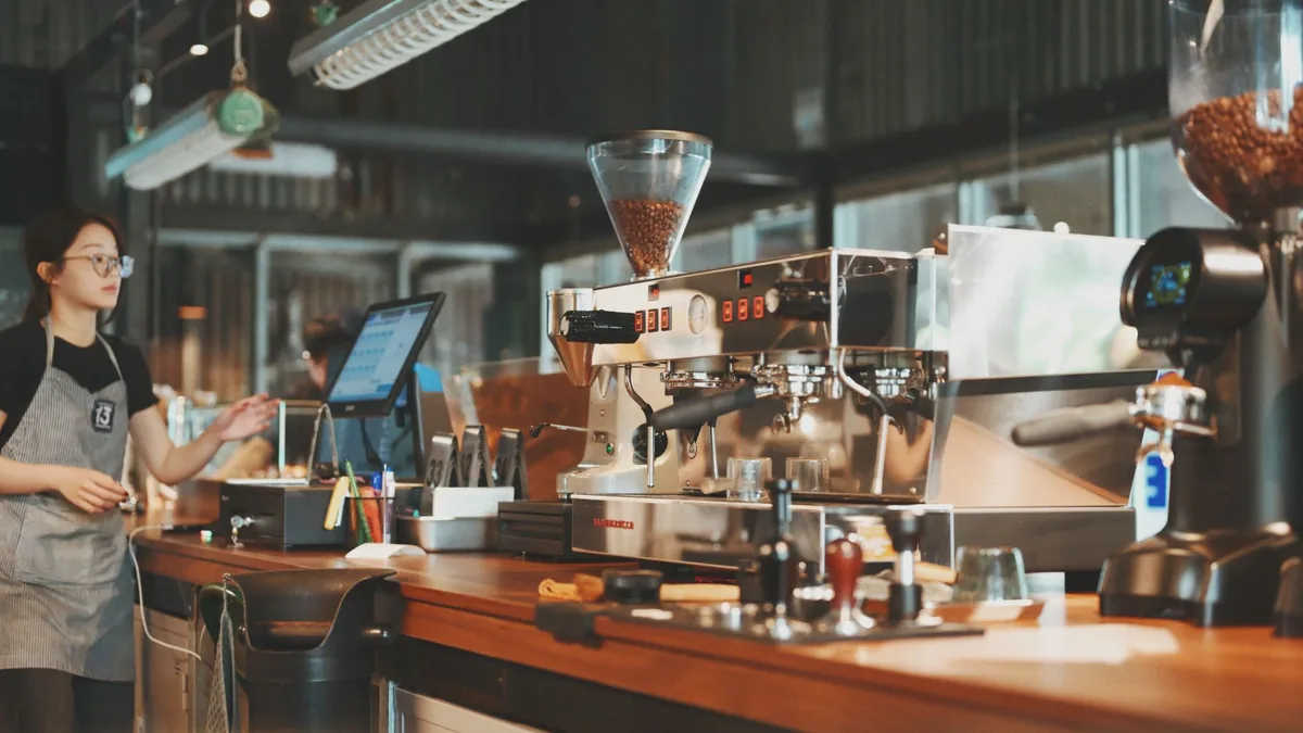 Barra de una cafetería con una máquina de espresso, molinillos y utensilios, y una barista trabajando al fondo.