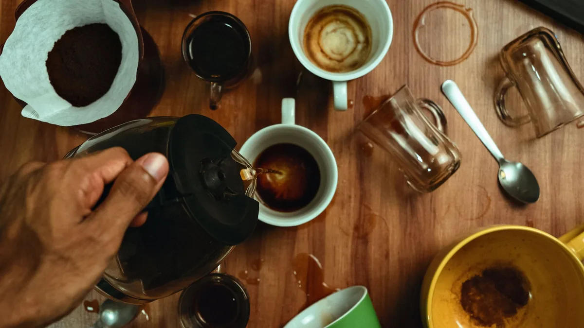 Vista cenital de una mesa de madera con varias tazas y vasos de café, un filtro de papel con café molido y una cucharilla, mientras una mano sirve café desde una jarra.