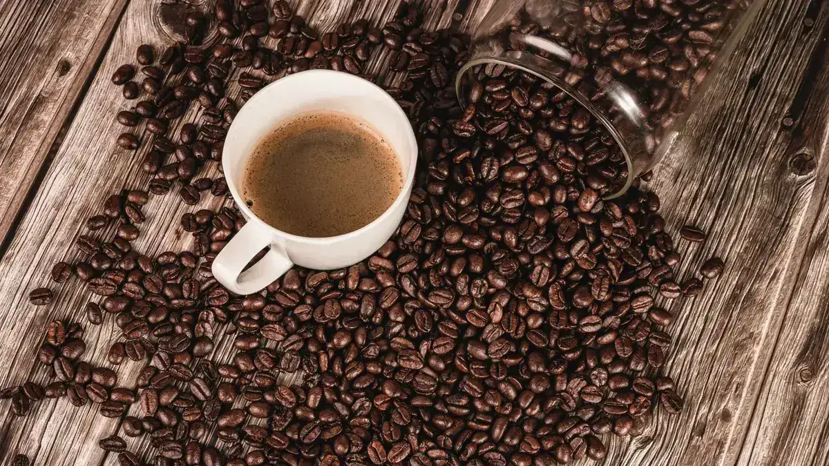 Taza blanca de café solo rodeada de granos de café tostado esparcidos sobre una mesa de madera rústica, con un tarro de cristal volcado al fondo.