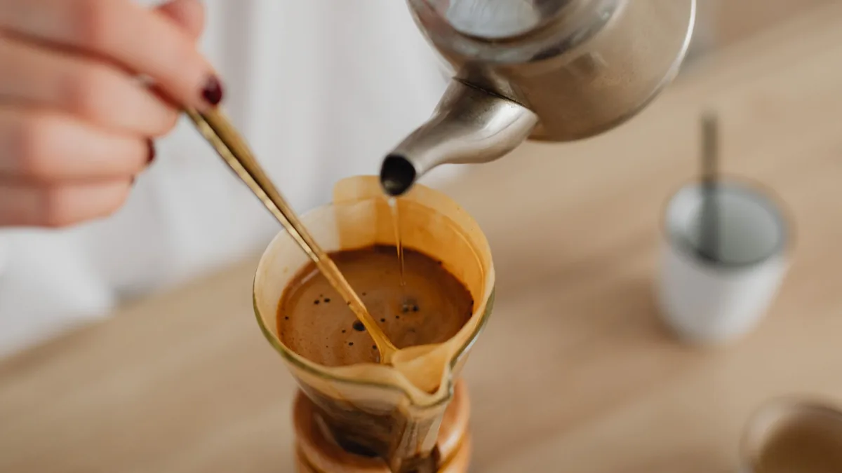 Una mano vierte agua caliente desde una tetera metálica sobre un filtro con café, mientras una cucharilla remueve la mezcla dentro de la jarra.