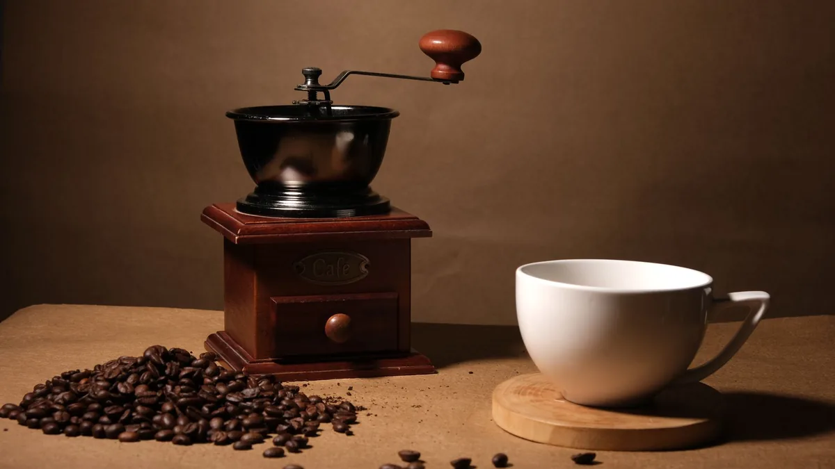 Molinillo de café manual antiguo de madera con manivela, junto a granos de café esparcidos y una taza blanca sobre un posavasos.