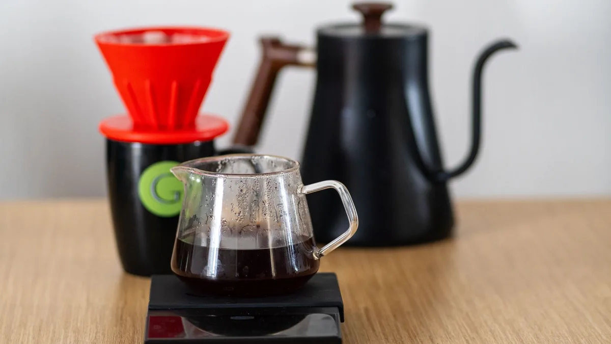 Cafetera de filtro V60 roja sobre una jarra de cristal con café recién hecho, apoyada en una báscula, con una tetera de cuello de cisne desenfocada al fondo.