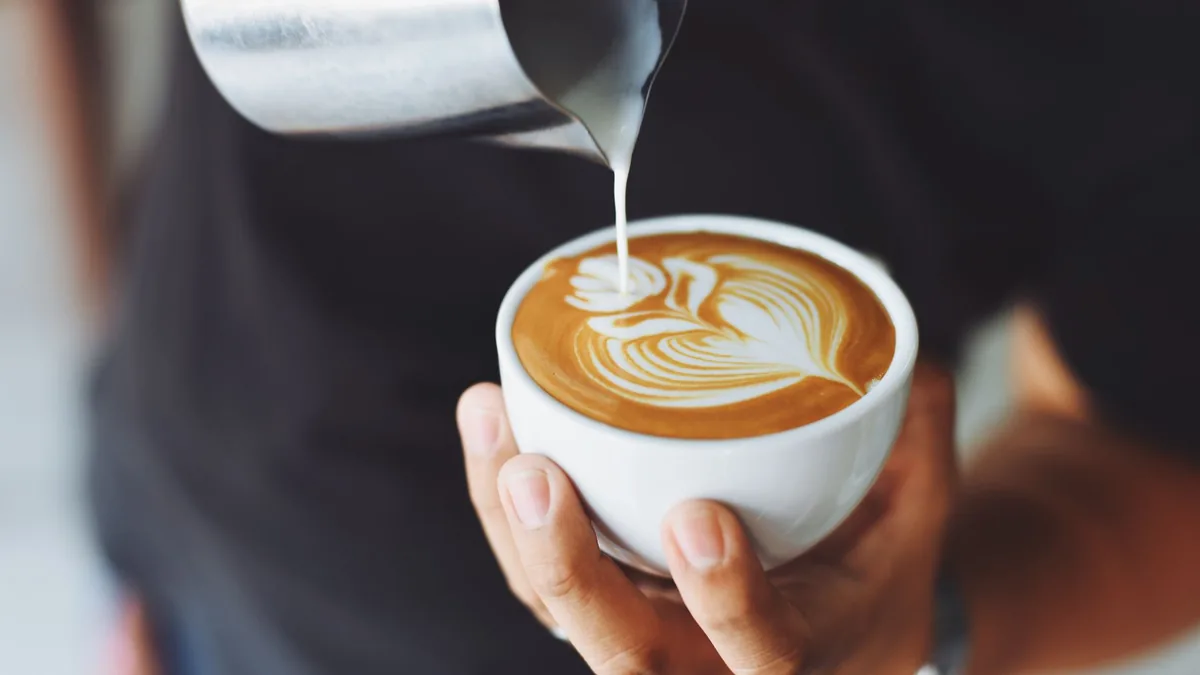 Una persona sostiene una taza blanca mientras se vierte leche desde una jarra metálica para dibujar una rosetta de latte art.